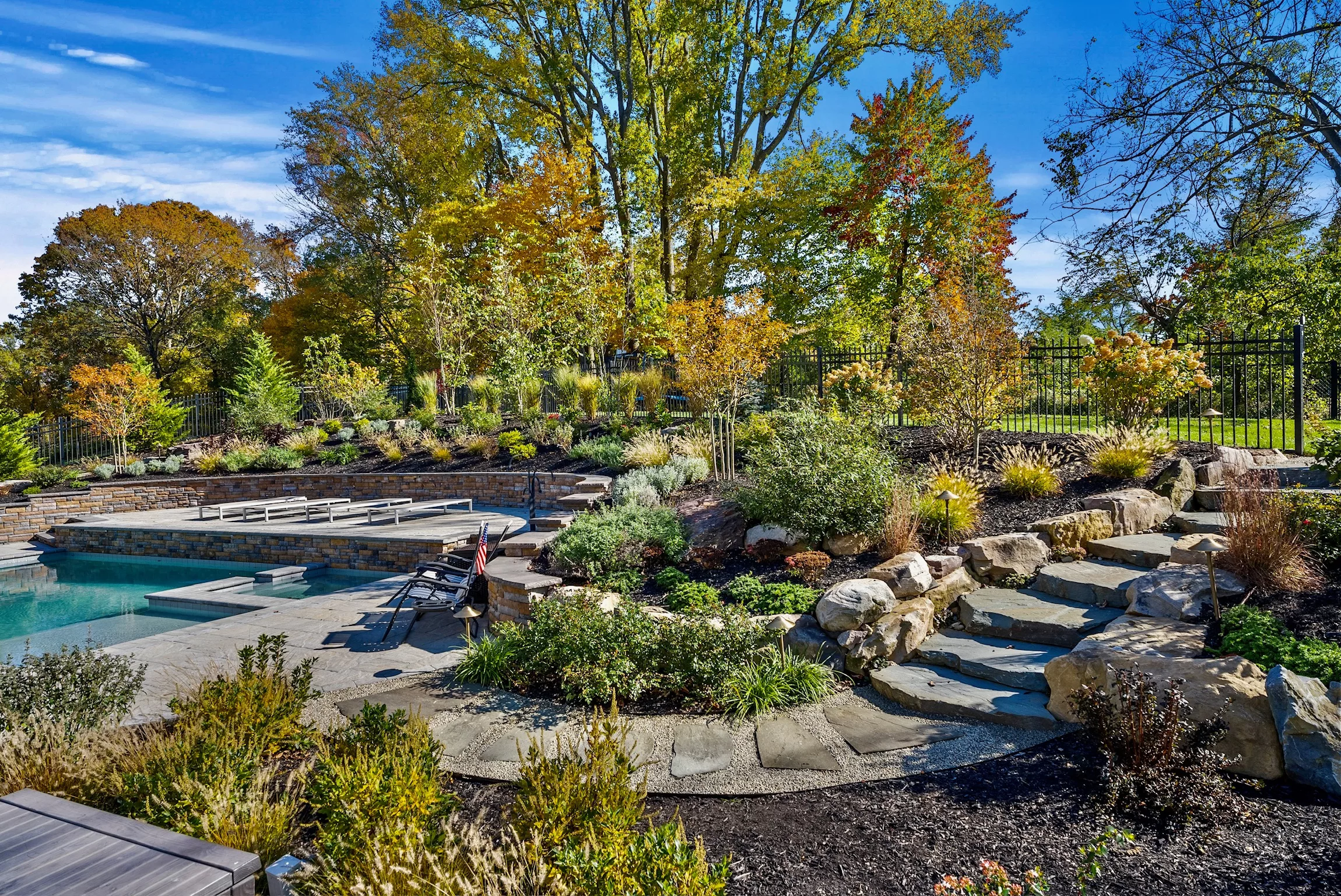 Stunning backyard pool surrounded by natural rock formations, landscaped garden beds, and a stone pathway under vibrant autumn trees.